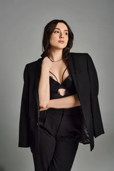 Una hermosa mujer de talla grande en un traje negro toma una postura confiada contra un fondo gris, exudando elegancia y estilo. - foto de stock