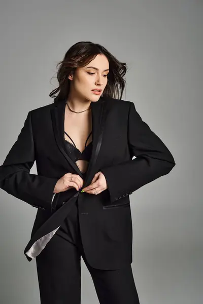 A beautiful plus-size woman confidently poses in a black suit and tie against a gray backdrop. — Stock Photo