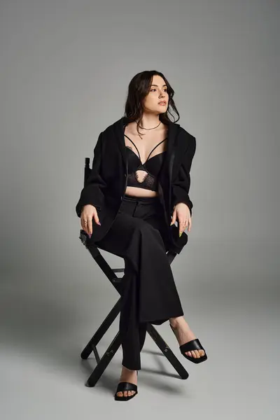 Una mujer de talla grande con un atuendo elegante sentada en una silla, cruzando sus piernas con gracia sobre un fondo gris. - foto de stock