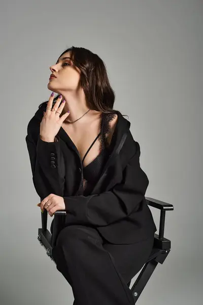 Una mujer de talla grande con un atuendo elegante se sienta en una silla, exudando confianza y encanto contra un fondo gris. - foto de stock