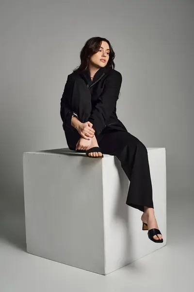 Una bella donna plus size in abiti eleganti siede elegantemente in cima a un blocco bianco su uno sfondo grigio. — Foto stock