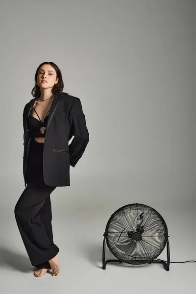 A beautiful plus size woman in a black suit standing gracefully next to a fan on a gray backdrop. — Photo de stock