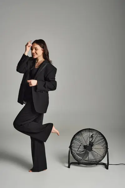 Una hermosa mujer de talla grande con un atuendo elegante, de pie con gracia delante de un ventilador, su traje revoloteando en la brisa. - foto de stock