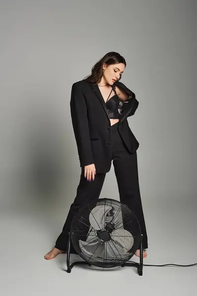 Stylish plus size woman in a suit standing confidently next to a fan on a gray backdrop, exuding poise and grace. — Stock Photo