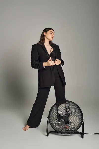 A stylish plus-size woman exudes confidence in a tailored suit, standing gracefully next to a spinning fan on a gray backdrop. — Stock Photo