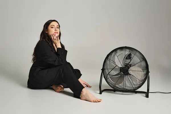 Une belle femme de taille plus en tenue élégante, assise gracieusement sur le sol à côté d'un ventilateur, sur fond gris. — Photo de stock