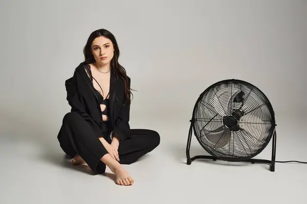 Une belle femme de grande taille en tenue élégante s'assoit gracieusement à côté d'un ventilateur tournant sur un fond gris. — Photo de stock