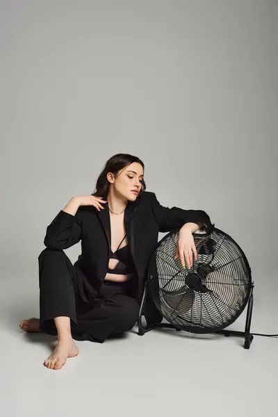 A beautiful plus size woman in stylish attire sitting peacefully on the floor next to a fan, enjoying the cool air. — Photo de stock