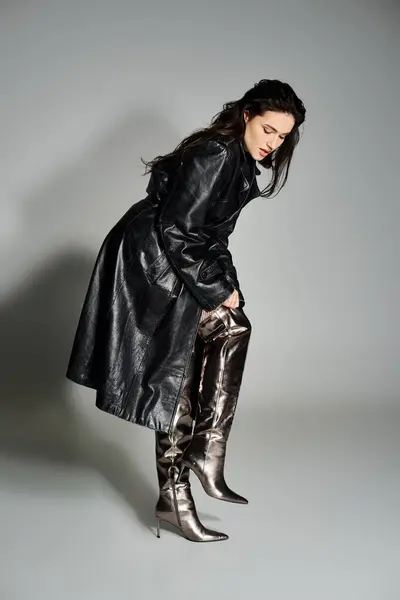 Uma bela mulher plus size posa em um casaco preto elegante e botas contra um pano de fundo cinza. — Fotografia de Stock