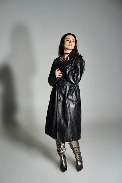 Приголомшлива жінка плюс розмір вдарив позу в шикарне чорне пальто і чоботи на сірому тлі. — стокове фото