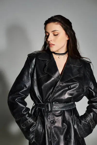 Красивая плюс размер женщина наносит уверенную позу в стильной черной кожаной траншеи пальто на нейтральном сером фоне. — стоковое фото