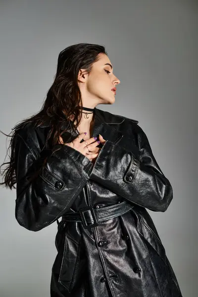 Una hermosa mujer de talla grande con un abrigo de cuero negro golpea una pose elegante contra un fondo gris. - foto de stock