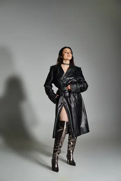 Uma mulher deslumbrante plus size exala glamour em um casaco preto e botas de coxa alta contra um pano de fundo cinza. — Fotografia de Stock