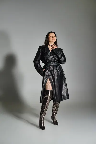 Una mujer elegante de talla grande se coloca con confianza en un abrigo negro y botas contra un telón de fondo gris, exudando elegancia. - foto de stock
