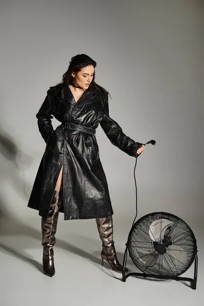 Plus la femme de taille respire la sophistication dans le trench coat, mettant en valeur le ventilateur dans une pose frappante sur fond gris. — Photo de stock