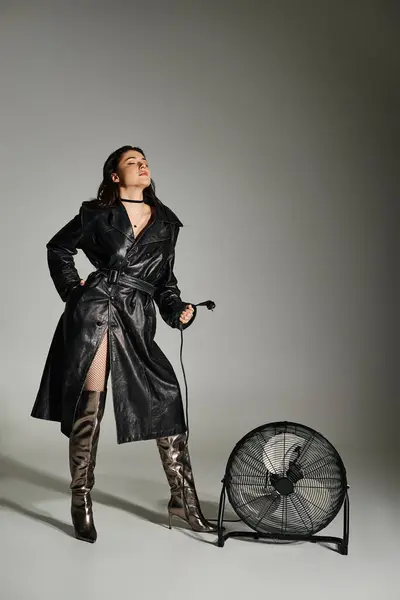 Hermosa mujer de talla grande en un abrigo negro de pie con gracia junto a un ventilador en un fondo gris. - foto de stock