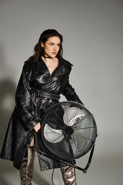 Une belle femme de taille plus posant dans un manteau en cuir à la mode, tenant un ventilateur, respirant la confiance et le style sur un fond gris. — Photo de stock