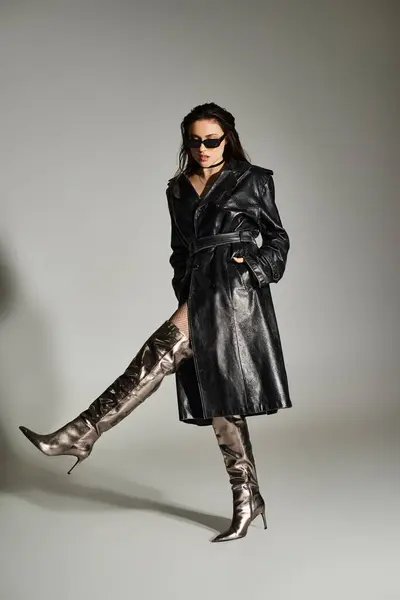 Una hermosa mujer de talla grande posa con confianza en un elegante abrigo negro y botas a juego contra un fondo gris. - foto de stock