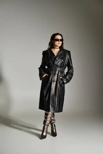 Une belle femme de taille plus respire la confiance dans un trench coat noir et des bottes sur fond gris. — Photo de stock