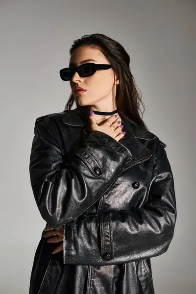 Una hermosa mujer de talla grande posa con confianza, vistiendo una chaqueta de cuero negro y gafas de sol sobre un fondo gris. - foto de stock