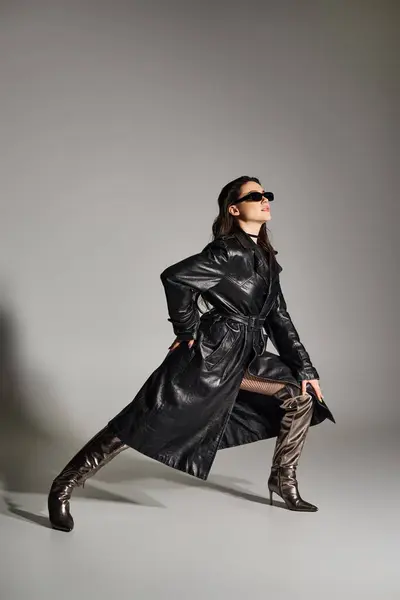Una hermosa mujer de talla grande posa con confianza en un abrigo negro y botas contra un telón de fondo gris, exudando estilo y gracia. - foto de stock