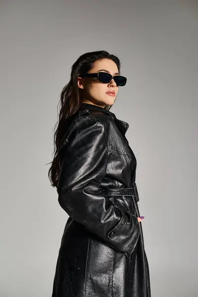 A beautiful plus size woman confidently poses in a black leather coat and stylish sunglasses against a gray backdrop. — Stock Photo