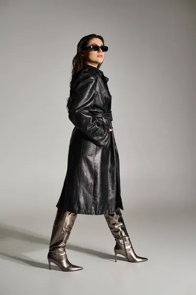 Elegante mujer de talla grande pavoneándose con confianza en un abrigo de cuero negro y botas contra un fondo gris llamativo. - foto de stock