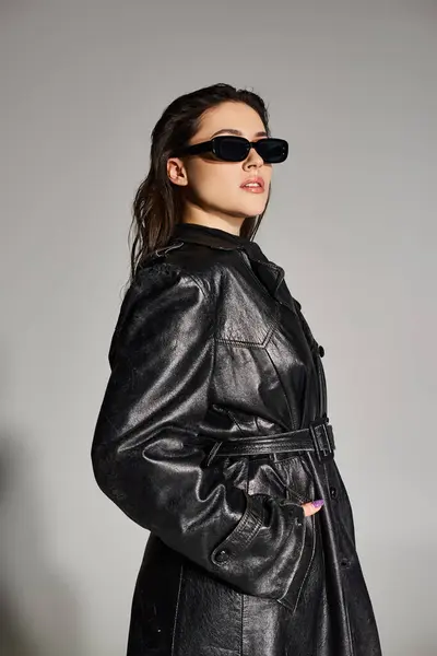Una hermosa mujer de talla grande posa en un elegante abrigo de cuero negro y gafas de sol sobre un fondo gris. - foto de stock