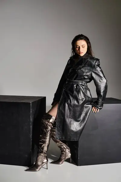 Una mujer de talla grande con un abrigo negro y botas apoyadas en una caja negra sobre un fondo gris, exudando elegancia y confianza. - foto de stock