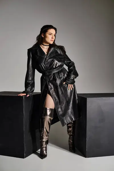 A stunning plus-size woman showcasing her style in a black trench coat and boots against a grey backdrop. — Stock Photo