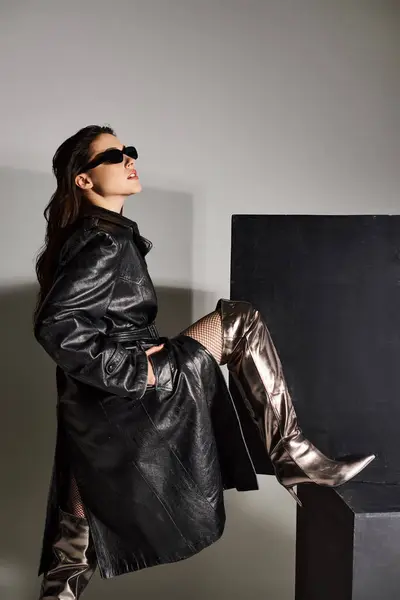 Una hermosa mujer de talla grande posa confiadamente con un abrigo negro y botas plateadas sobre un fondo gris. - foto de stock