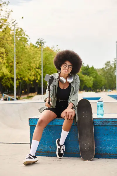 Молодая афроамериканка с вьющимися волосами сидит в скейт-парке, держа в руках селфи-палку. — стоковое фото
