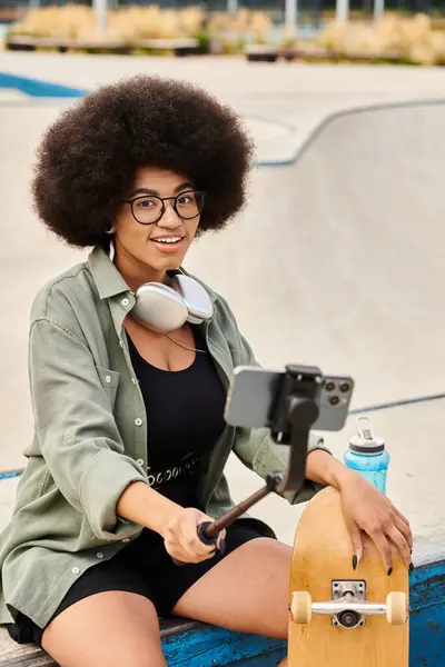 Joven mujer afroamericana con el pelo rizado sentado en una cornisa, sosteniendo un palo selfie en un parque de skate vibrante. - foto de stock