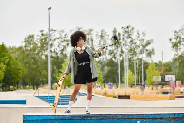An African American woman with curly hair confidently holds a skateboard in a vibrant outdoor skate park. — Stock Photo