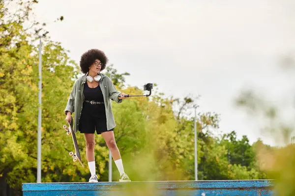 Junge Afroamerikanerin mit lockigem Haar hält selbstbewusst Skateboard und Selfie-Stick in der Hand — Stockfoto