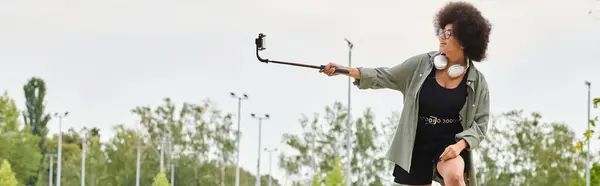 Junge Afroamerikanerin mit Kopfhörern und lockigem Haar beim Skateboarden, während sie einen Stock in einem Skatepark hält. — Stockfoto