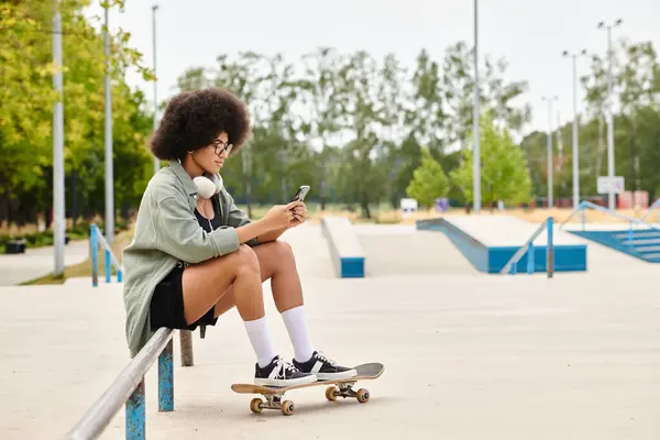 Une jeune Afro-Américaine aux cheveux bouclés faisant une pause en s'asseyant sur un banc avec sa planche à roulettes dans un skate park. — Photo de stock