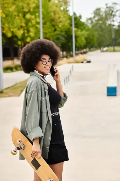 Une jeune afro-américaine aux cheveux bouclés multitâches en tenant une planche à roulettes et en parlant sur un téléphone portable dans un skate park. — Photo de stock