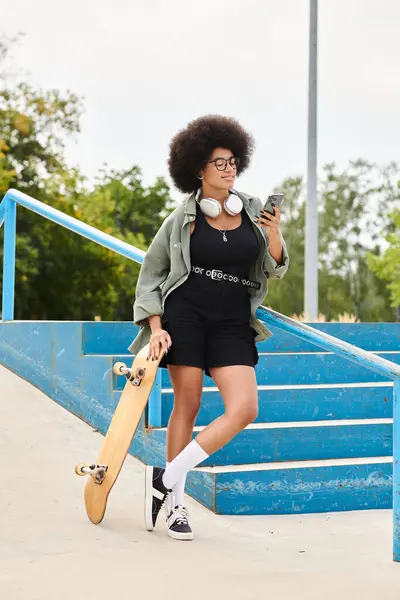 Una giovane donna afroamericana con i capelli ricci che tiene uno skateboard e un cellulare all'aperto in uno skate park. — Foto stock