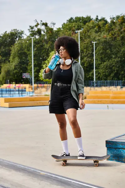 Молодая афроамериканка с вьющимися волосами уверенно стоит на скейтборде, путешествуя по яркому парку. — стоковое фото