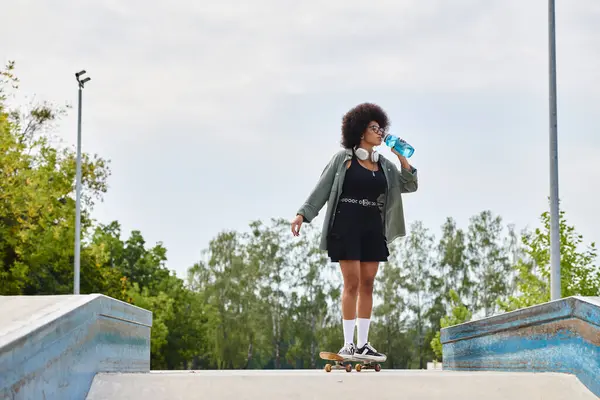 An African American woman with curly hair, standing on a skateboard, casually drinking water in a skate park. — Stock Photo
