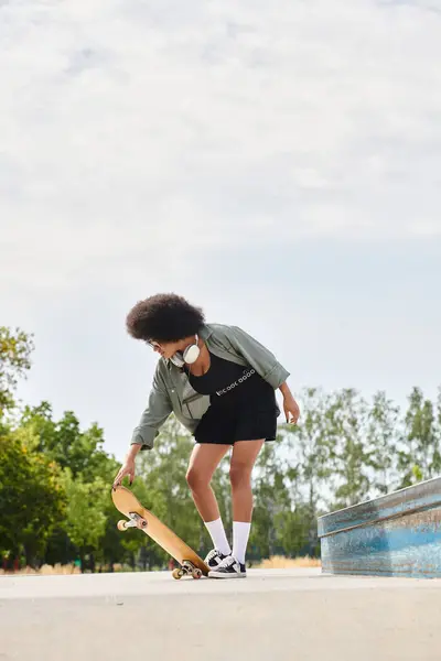 Mujer afroamericana con pelo rizado se desliza en monopatín en un elegante vestido negro en un parque de skate al aire libre. - foto de stock