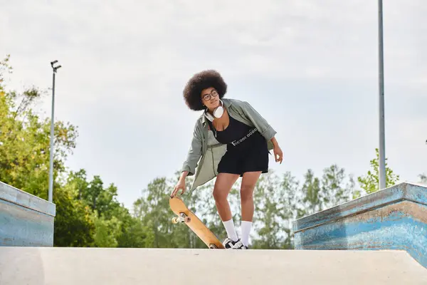 Una joven afroamericana con el pelo rizado patina con confianza en un elegante vestido negro en un vibrante parque de skate al aire libre. - foto de stock