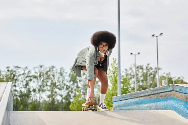 Joven mujer afroamericana con el pelo rizado hábilmente monta un monopatín en una rampa en un parque de skate al aire libre. - foto de stock