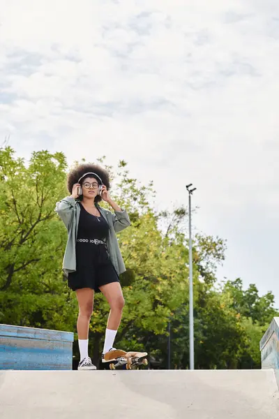 A young African American woman with curly hair confidently stands on top of a skateboard ramp in a skate park. — Stock Photo