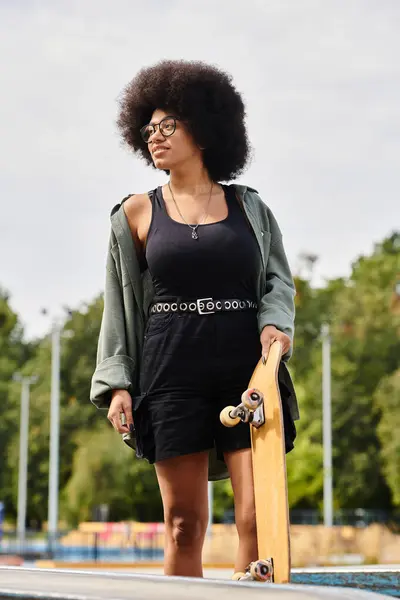 Una joven afroamericana sostiene con confianza un monopatín en la parte superior de una rampa en un parque de skate. - foto de stock