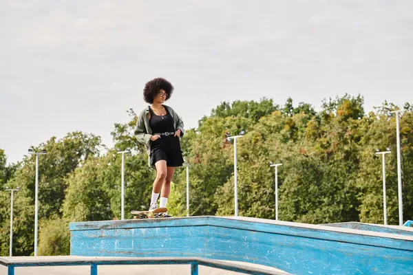 An African American woman with curly hair confidently stands on top of a skateboard ramp in an outdoor skate park. — Stock Photo