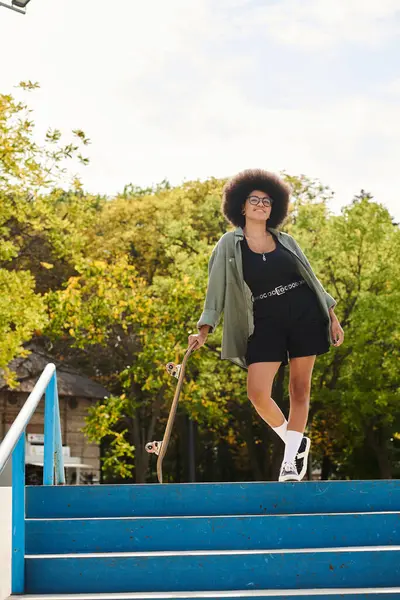 Une jeune afro-américaine aux cheveux bouclés monte en toute confiance un escalier tout en tenant une planche à roulettes. — Photo de stock