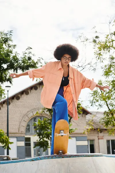 Una joven afroamericana con el pelo rizado monta sin miedo un monopatín al lado de una rampa en un parque de skate al aire libre. - foto de stock