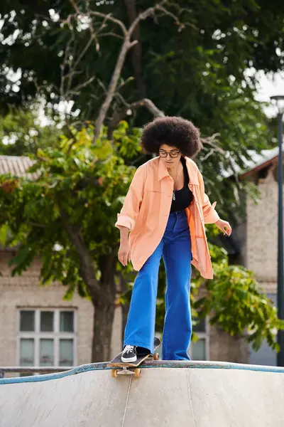 Una joven afroamericana con el pelo rizado monta hábilmente un monopatín en una rampa en un parque de skate al aire libre. - foto de stock